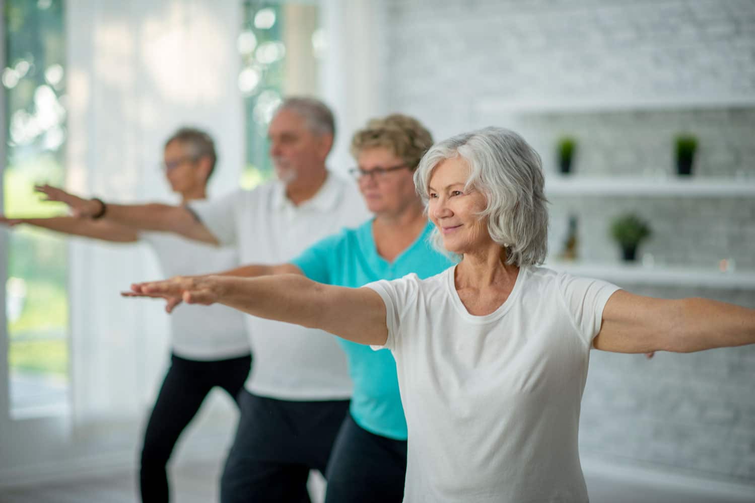 A small group of seniors stand evenly spready apart in a studio with their hands extended as they stretch and balance during a Tai Chi class. They are each dressed comfortably and are focused on the movements.