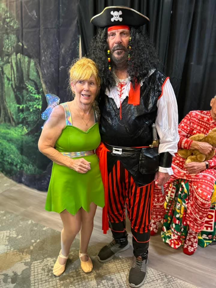 A resident dressed as Tinker Bell poses with another resident dressed as Captain Hook in a pirate costume, featuring a black hat with a skull emblem and red and black striped pants, standing in front of a Peter Pan themed backdrop.
