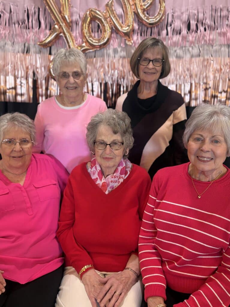 Residents in pink outfits posing together under a “Love” banner at the Villas at Stoney Point Galentine’s Day gathering.