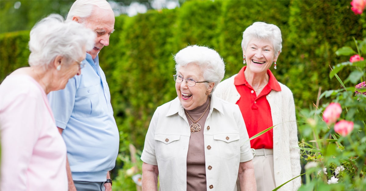 Group of older adults gathered outdoors in a garden setting, standing among greenery and flowers while interacting together in a landscaped courtyard.