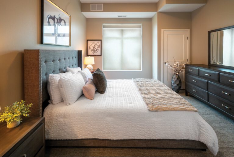 Apartment bedroom with bed, upholstered headboard, dresser, nightstands, and window in a comfortable living space at Villas at Stoney Point