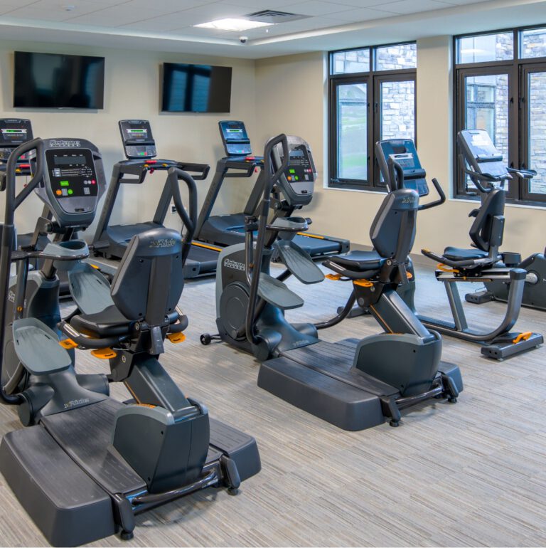 Fitness room with treadmills, recumbent bikes, elliptical machines, wall-mounted televisions, and large windows at Villas at Stoney Point