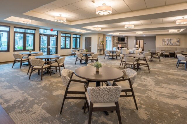 Spacious community room with multiple round tables, upholstered chairs, large windows, and a fireplace seating area at Villas at Stoney Point