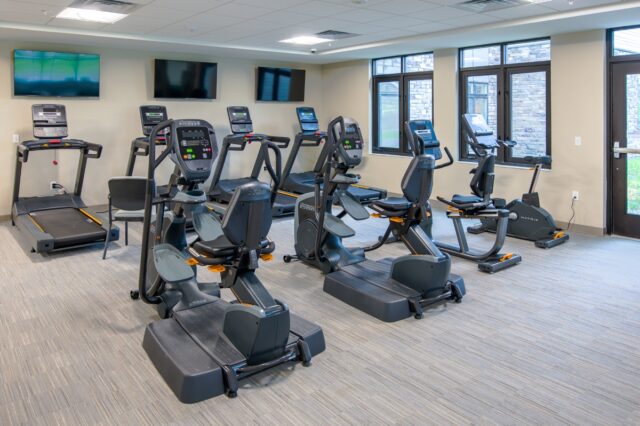 Fitness center with treadmills, recumbent bikes, elliptical machines, wall-mounted televisions, and large windows at Villas at Stoney Point