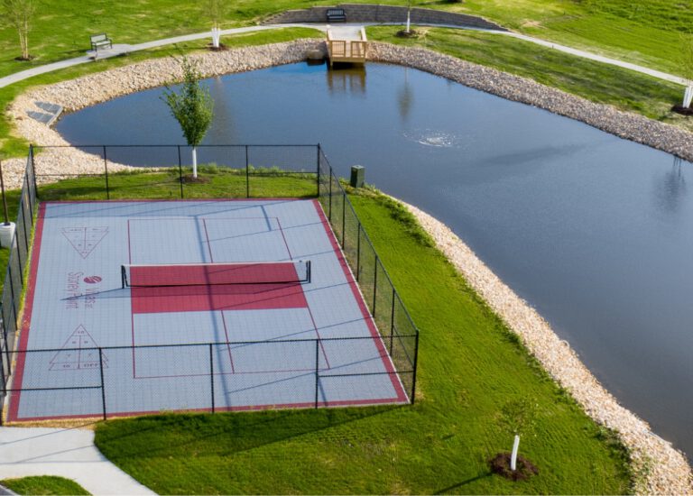 Fenced outdoor pickleball court beside a landscaped pond with walking paths and seating areas at Villas at Stoney Point