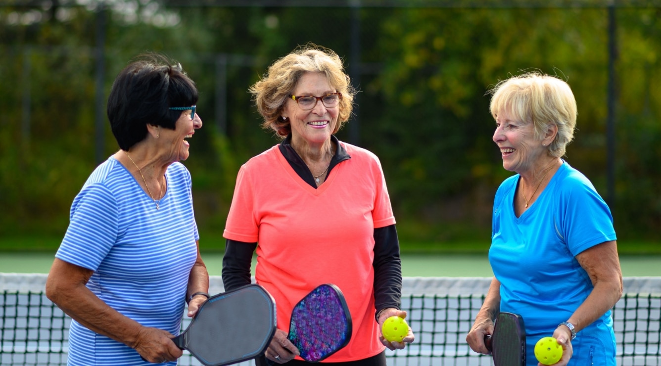 Three older adults holding pickleball paddles, while standing on an outdoor court, representing active social lifestyle like Villas at Stoney Point