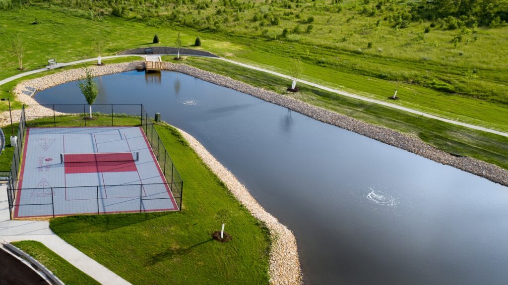 Aerial view of a pond with surrounding walking paths and a fenced pickleball court at Villas at Stoney Point