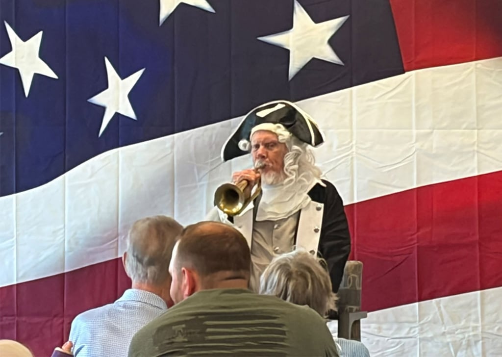 Resident dressed as a country music performer with braided hair and a red bandana holds a microphone in front of an American flag backdrop during the America 250 celebration at Villas at Stoney Point.